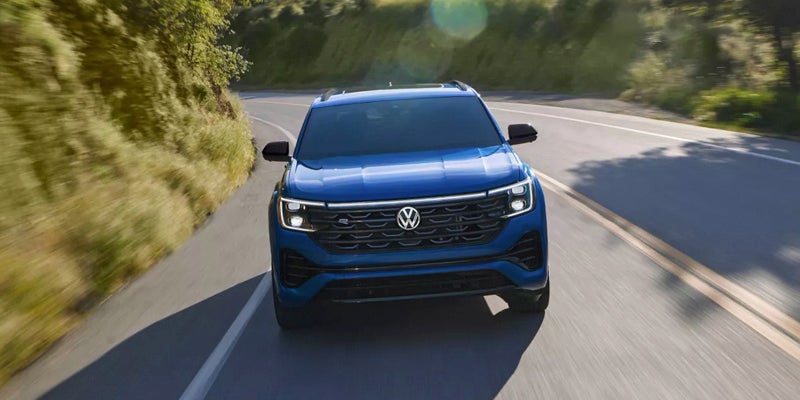 A blue VW Atlas Cross Sport SUV drives towards the viewer on a winding road with green hills.